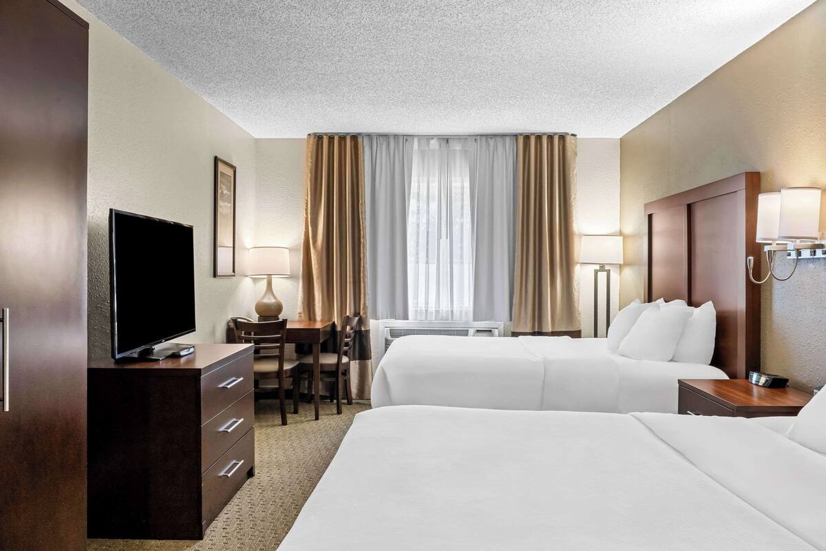 Ein Hotelzimmer mit zwei Betten, einem Schreibtisch mit Stuhl, einem Fernseher und einem Fenster mit Vorhängen.