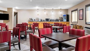 A hotel dining area with red upholstered chairs and tables, a kitchen in the background, and a flat-screen TV mounted on the wall. - Comfort Inn Worland Hwy 16 to Yellowstone (Worland)
