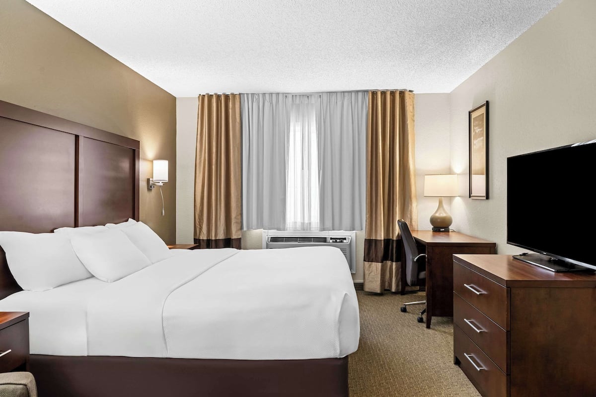 Ein Hotelzimmer mit einem großen Bett, einem Schreibtisch mit Computer, einem Fernseher und einem Fenster mit Vorhängen.