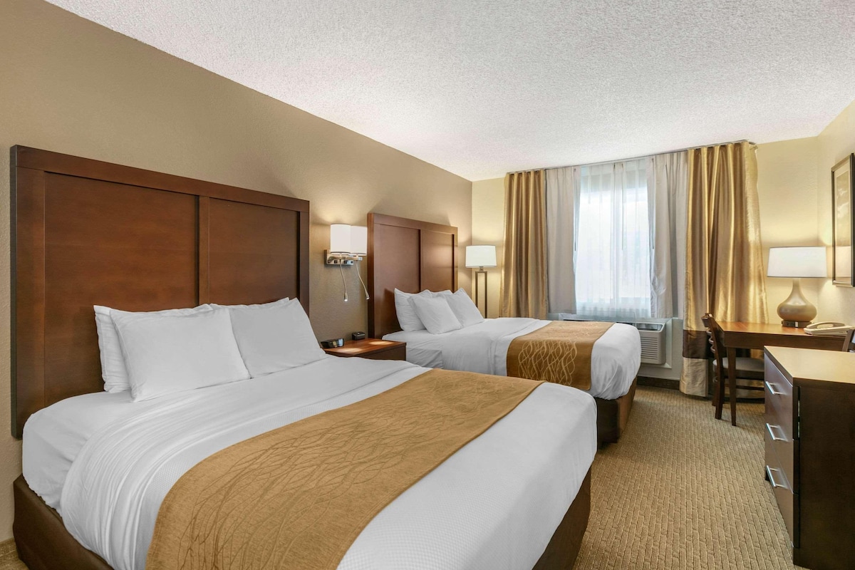 A hotel room with two beds, a desk, a chair, and a lamp.