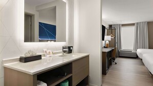 Une chambre d’hôtel avec un grand miroir, un lavabo, un bureau avec une lampe, un lit et une fenêtre avec des rideaux.