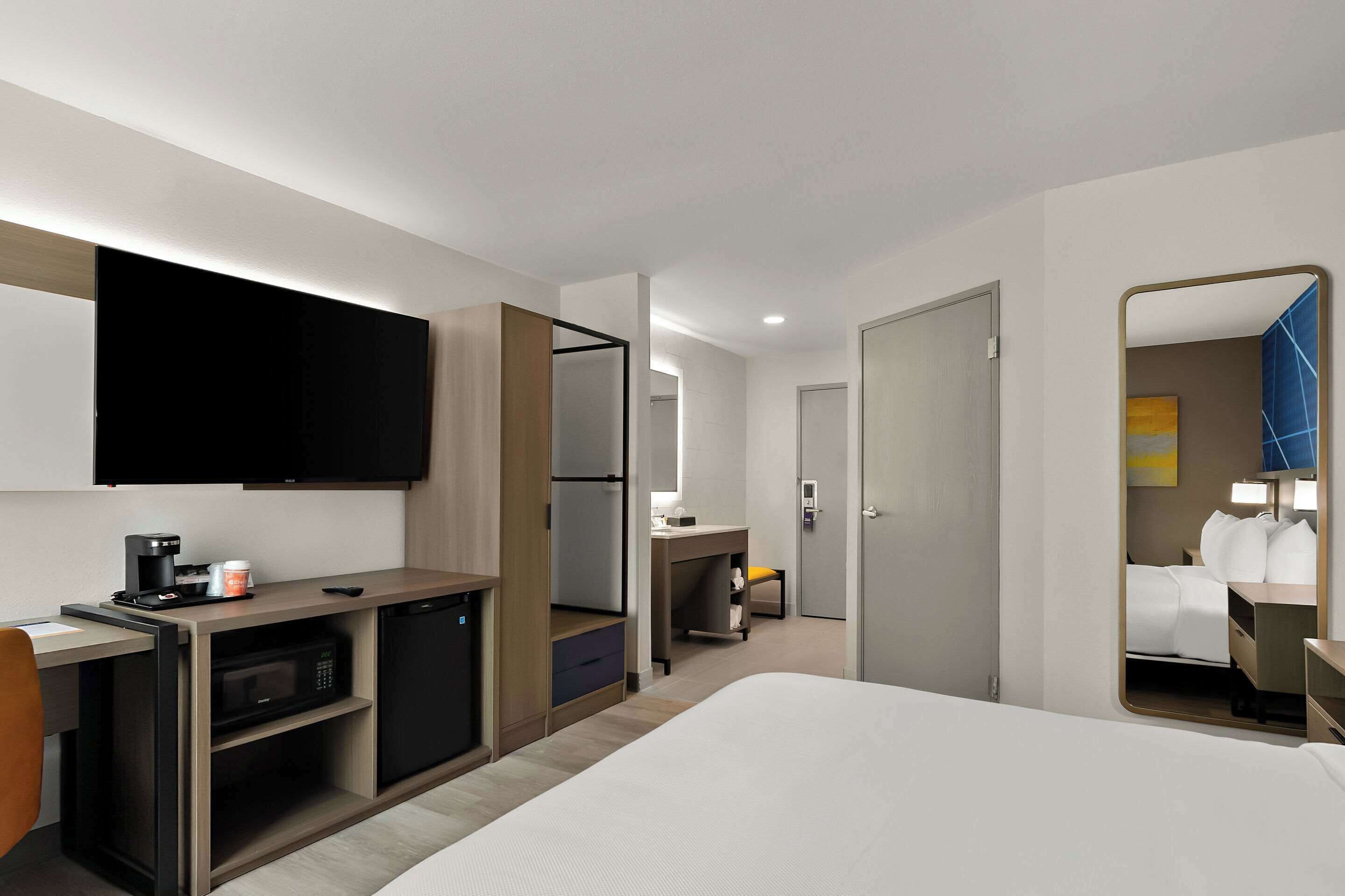 Een hotelkamer met een bed, een bureau met een magnetron, een koffiezetapparaat, een televisie, een spiegel en een deur naar een badkamer.