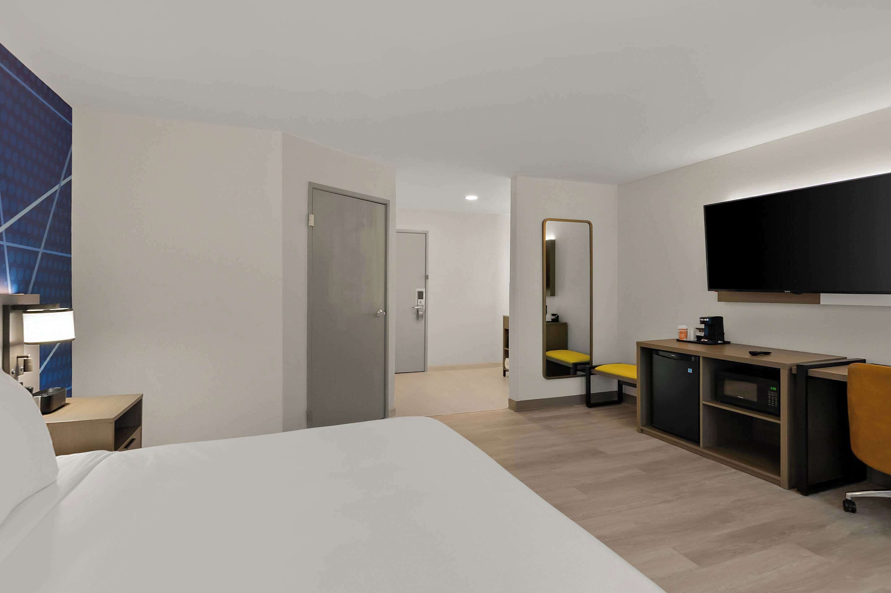 A hotel room with a bed, a desk with a computer, a TV, a mirror, and a door leading to another room.