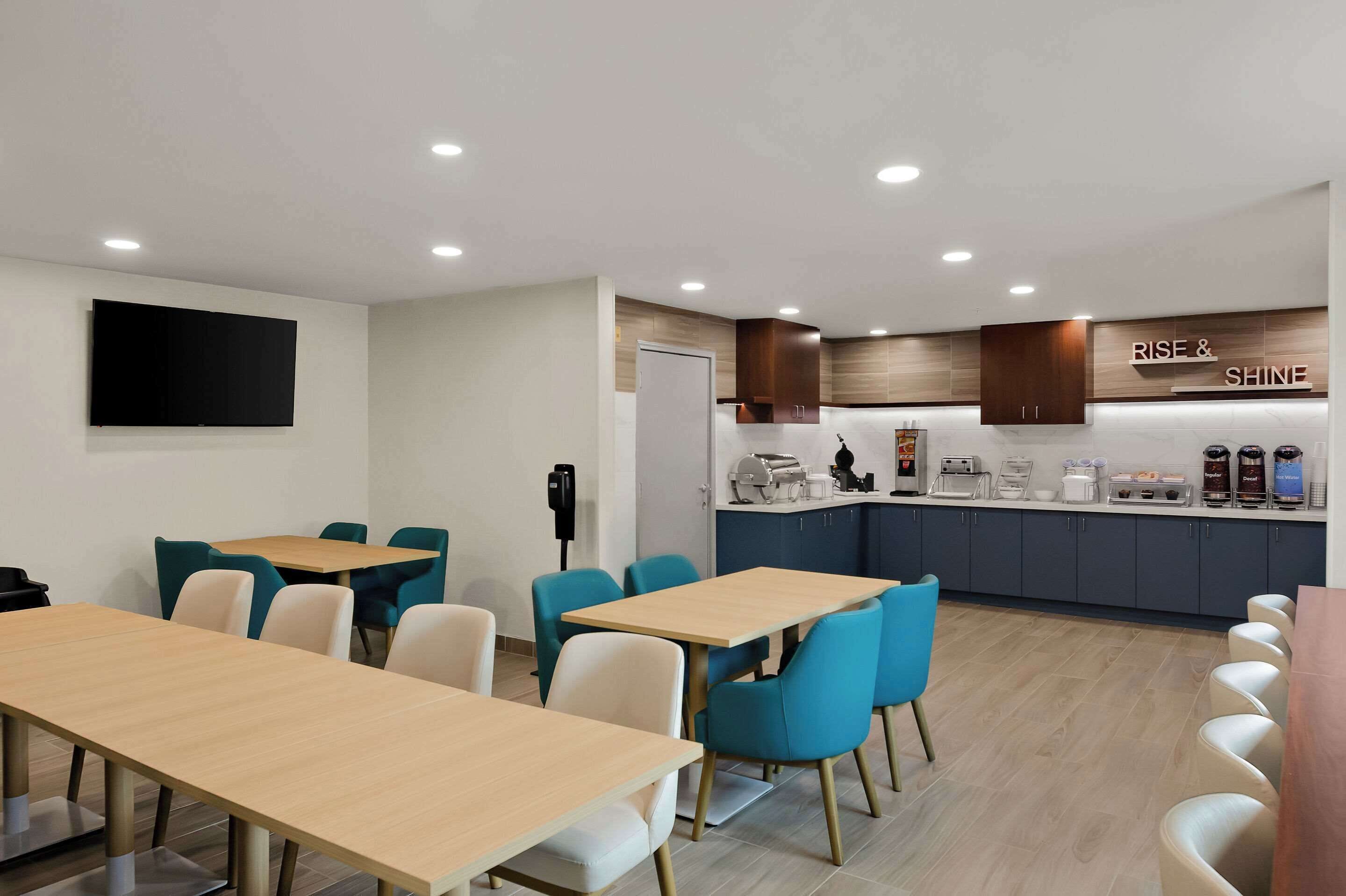 Un comedor moderno con mesas de madera y sillas azules, una cocina con un letrero que dice 'RISE & SHINE' y un televisor de pantalla plana montado en la pared.