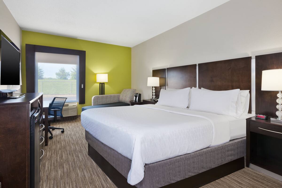 Ein Hotelzimmer mit einem großen Bett, einem Schreibtisch mit Flachbildfernseher, einem Sessel und einer Schreibtischlampe.