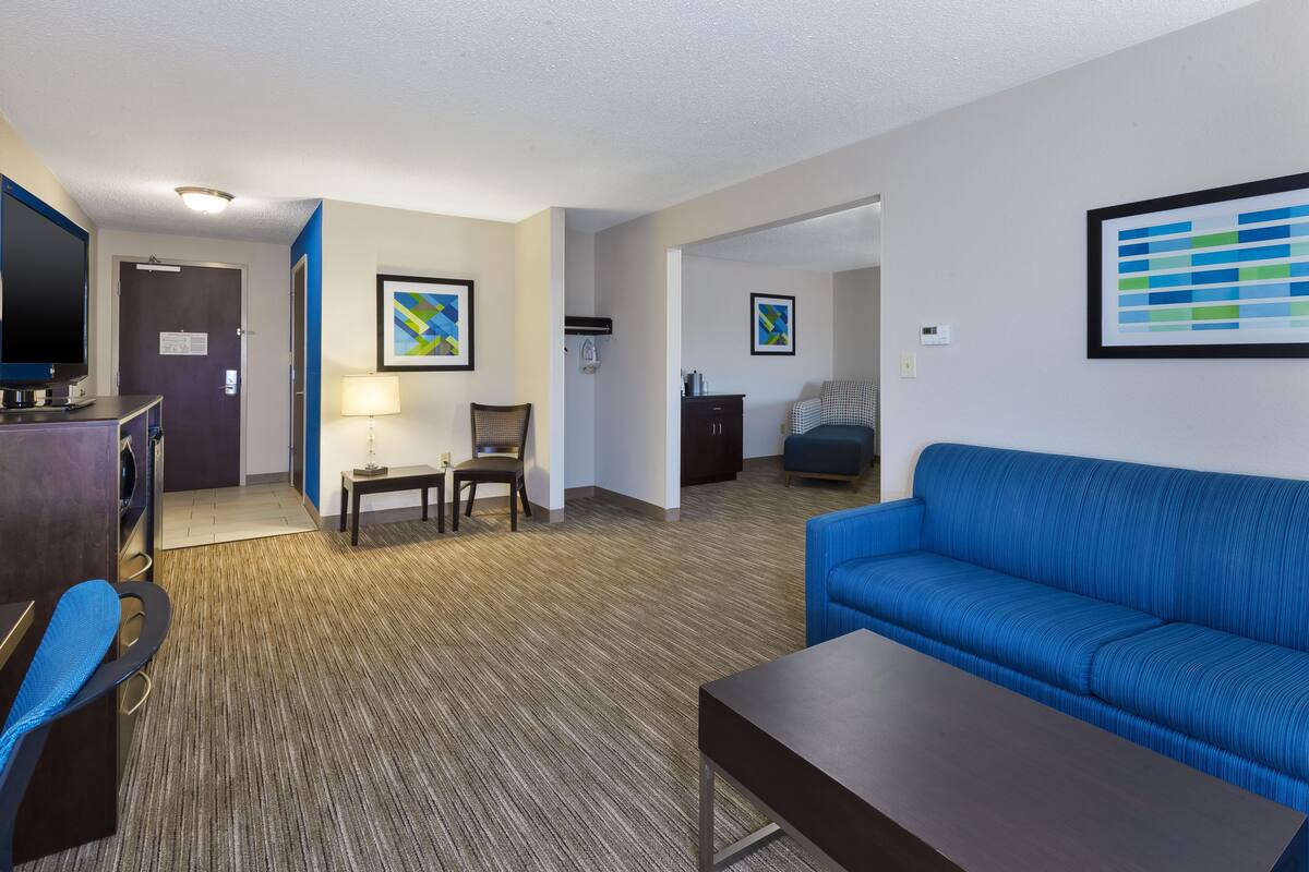 Ein Hotelzimmer mit blauem Sofa, einem kleinen Tisch, einem Stuhl, einem Schreibtisch mit Computer und einem Fernseher. An den Wänden befinden sich zwei gerahmte Kunstwerke und eine Tür zu einem weiteren Raum.