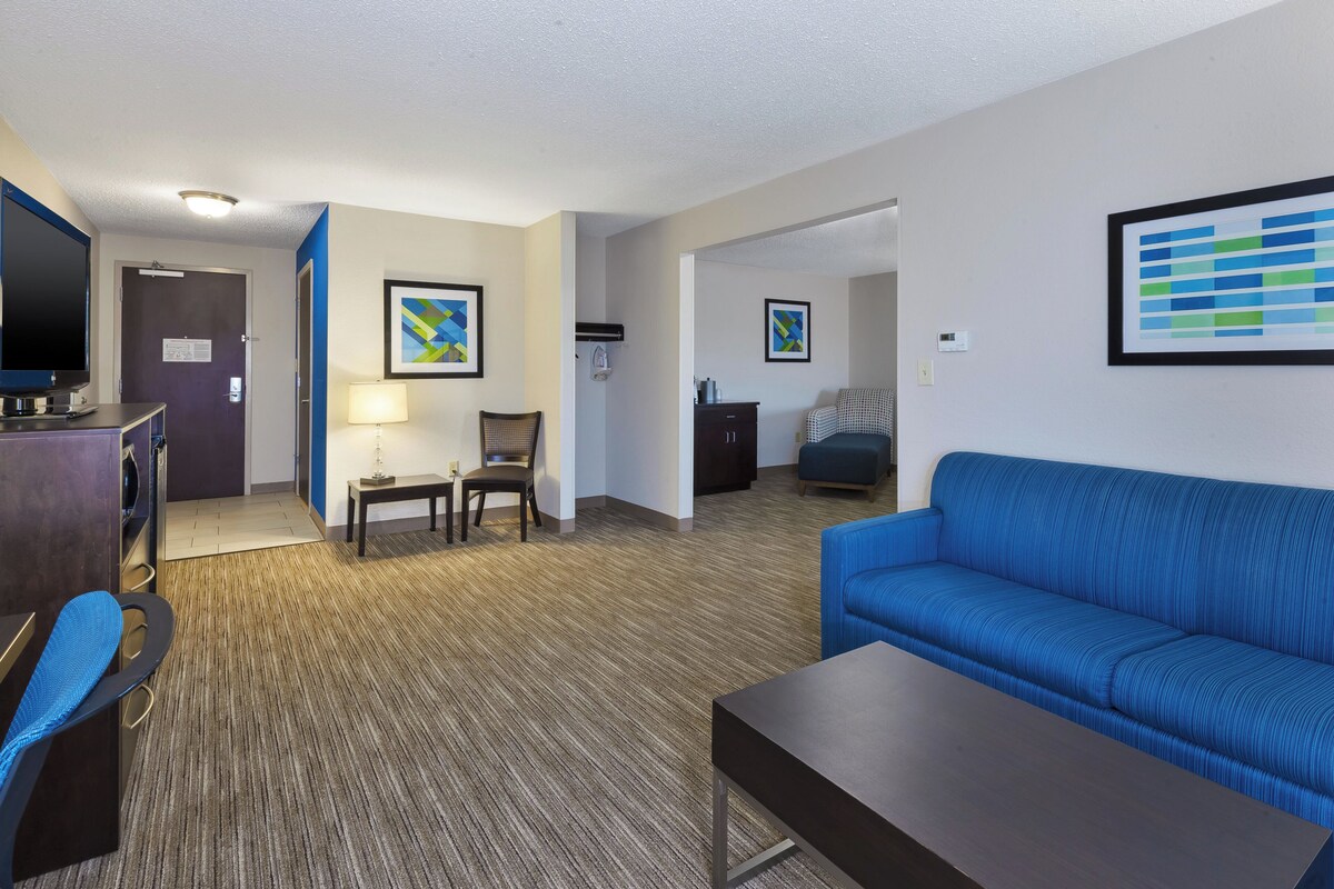 A hotel room with a blue sofa, a small table, a chair, a desk with a computer, and a TV. There are two framed artworks on the walls and a door leading to another room.