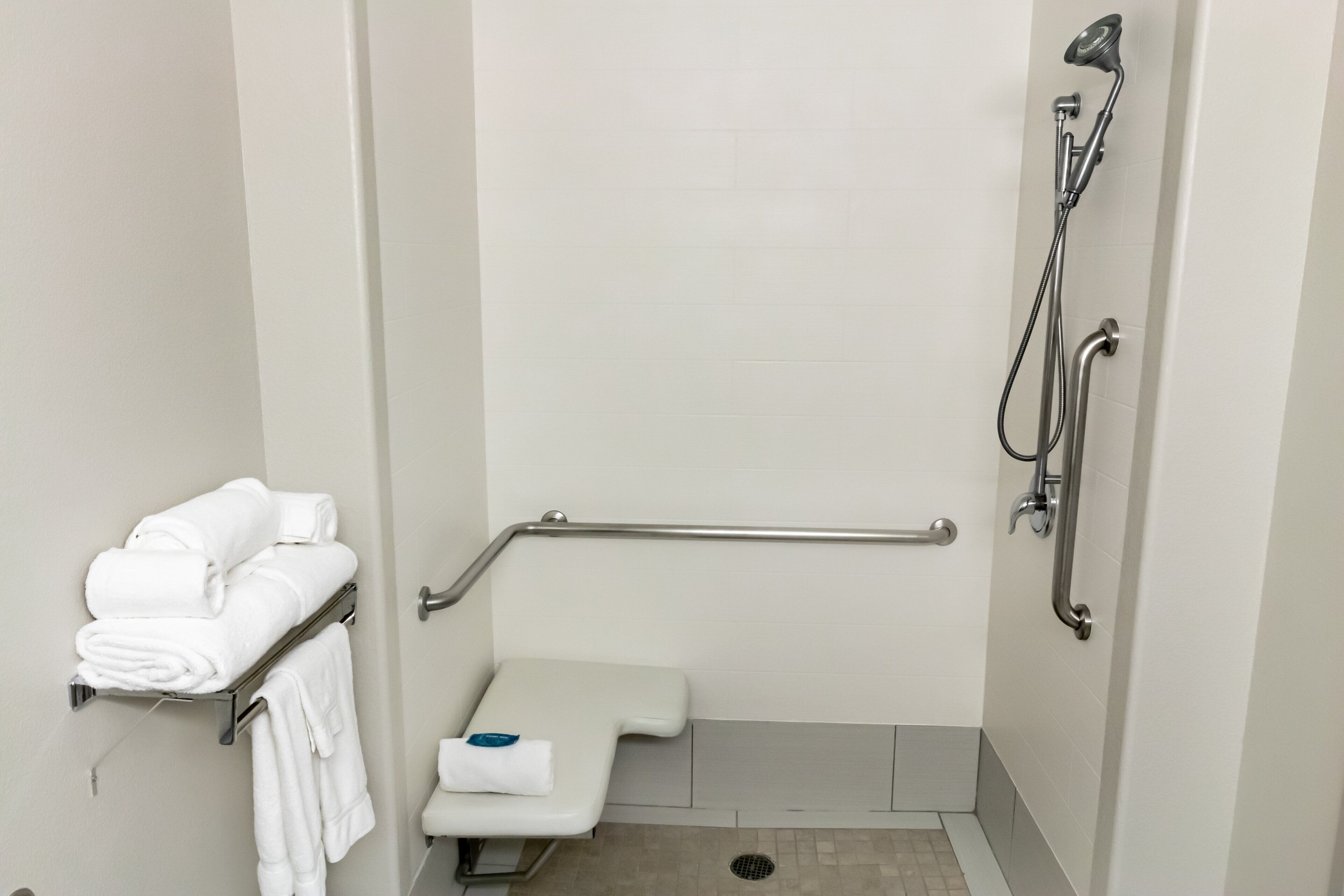 A wheelchair-accessible shower stall with a grab bar, a toilet with a safety bar, and a towel rack with folded towels.