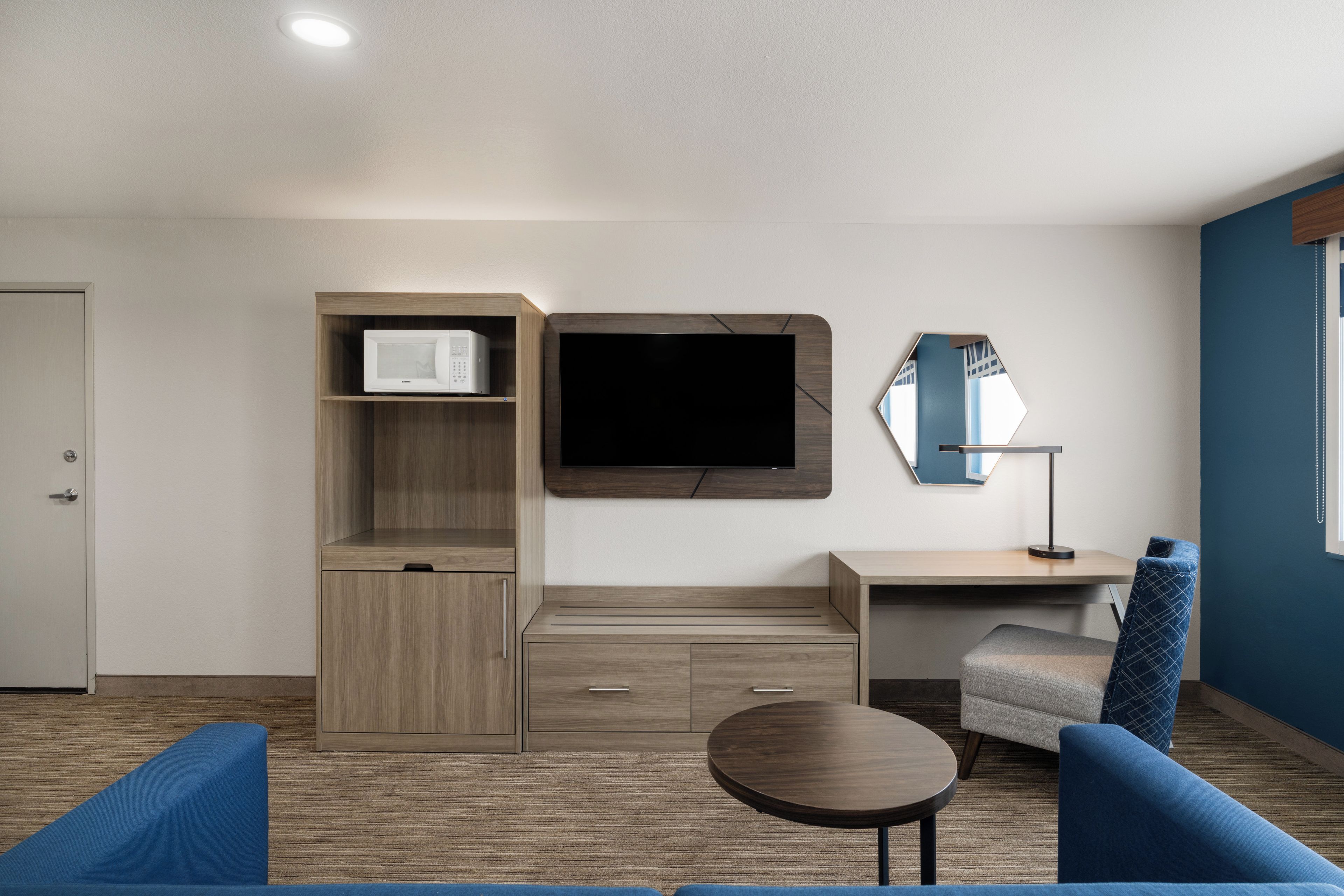 Quarto moderno com televisão de tela plana na parede, uma pequena escrivaninha com cadeira, micro-ondas em armário e sofá azul.