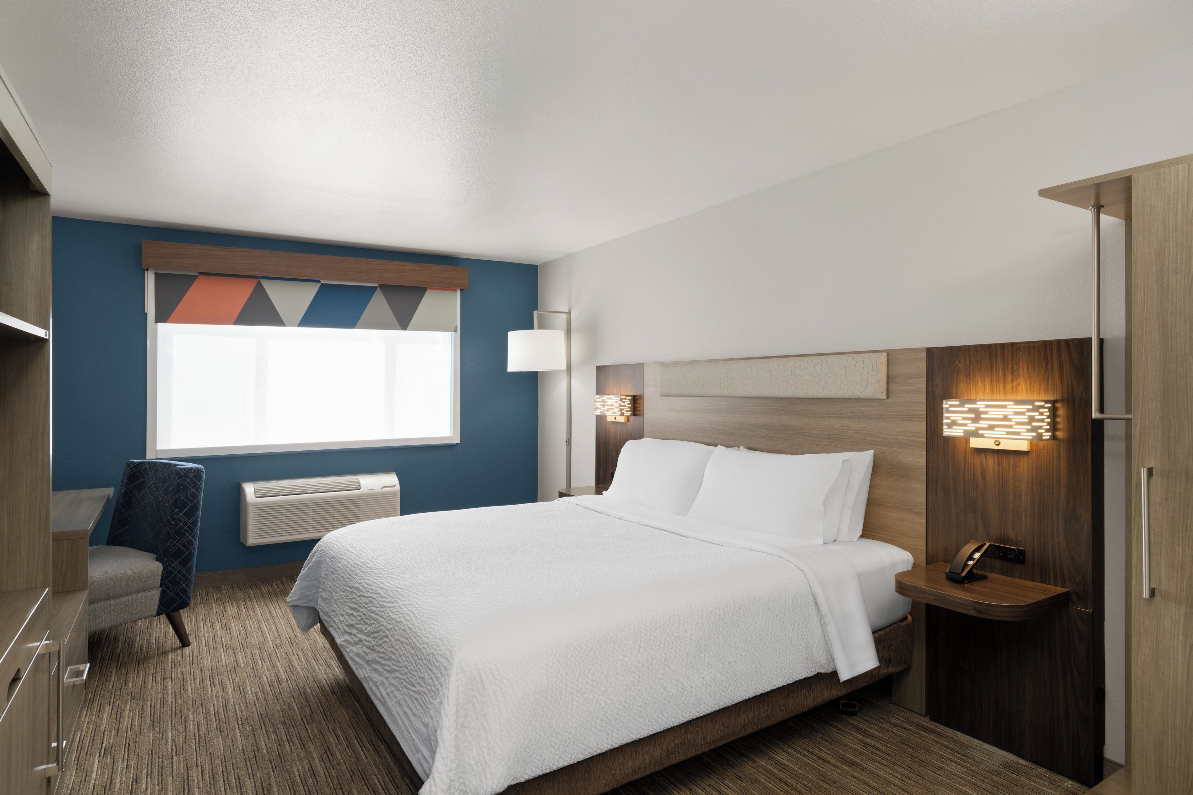 A hotel room with a bed, a desk, a chair, a nightstand, and a wall-mounted lamp.