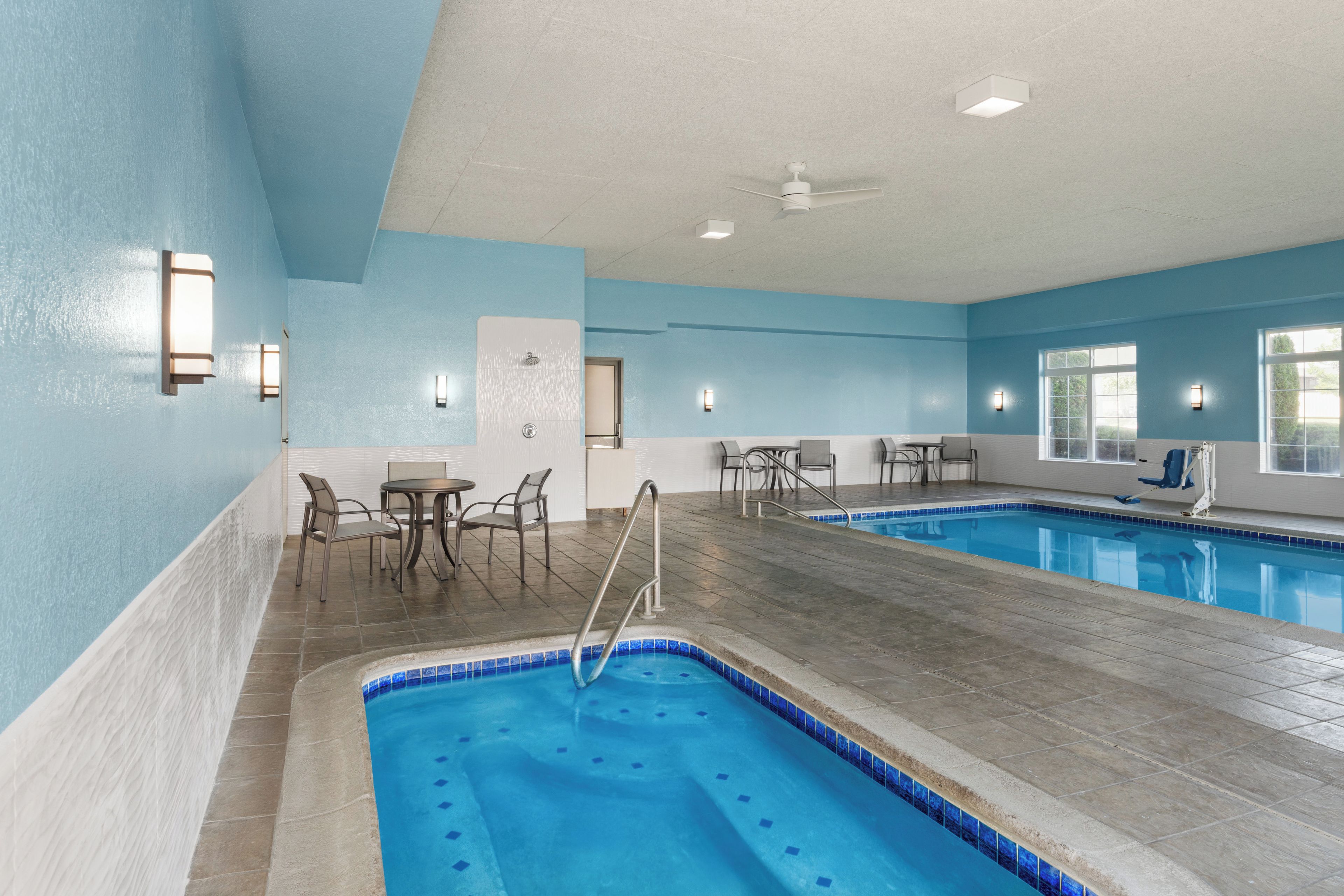 An indoor swimming pool with a blue tiled edge, a ceiling fan, and seating areas with tables and chairs.