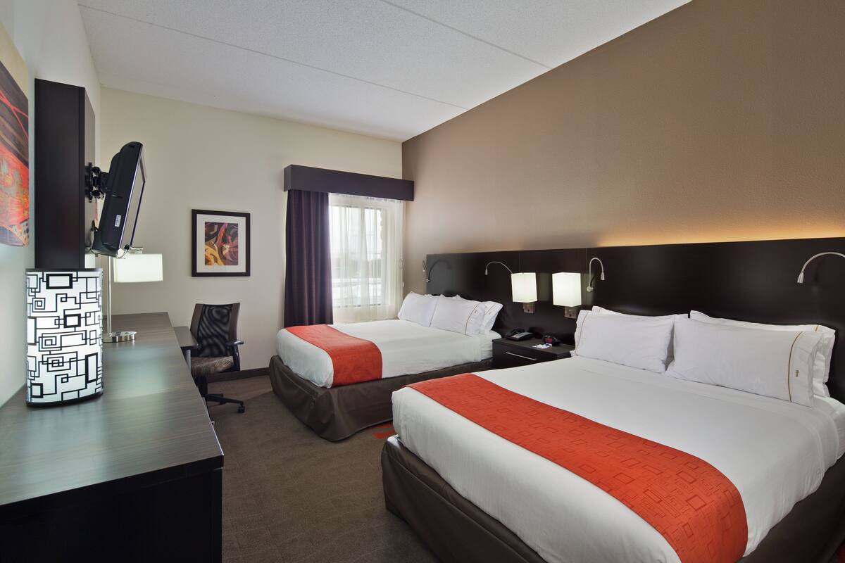 A hotel room with two beds, a desk, a chair, a lamp, and a painting on the wall.