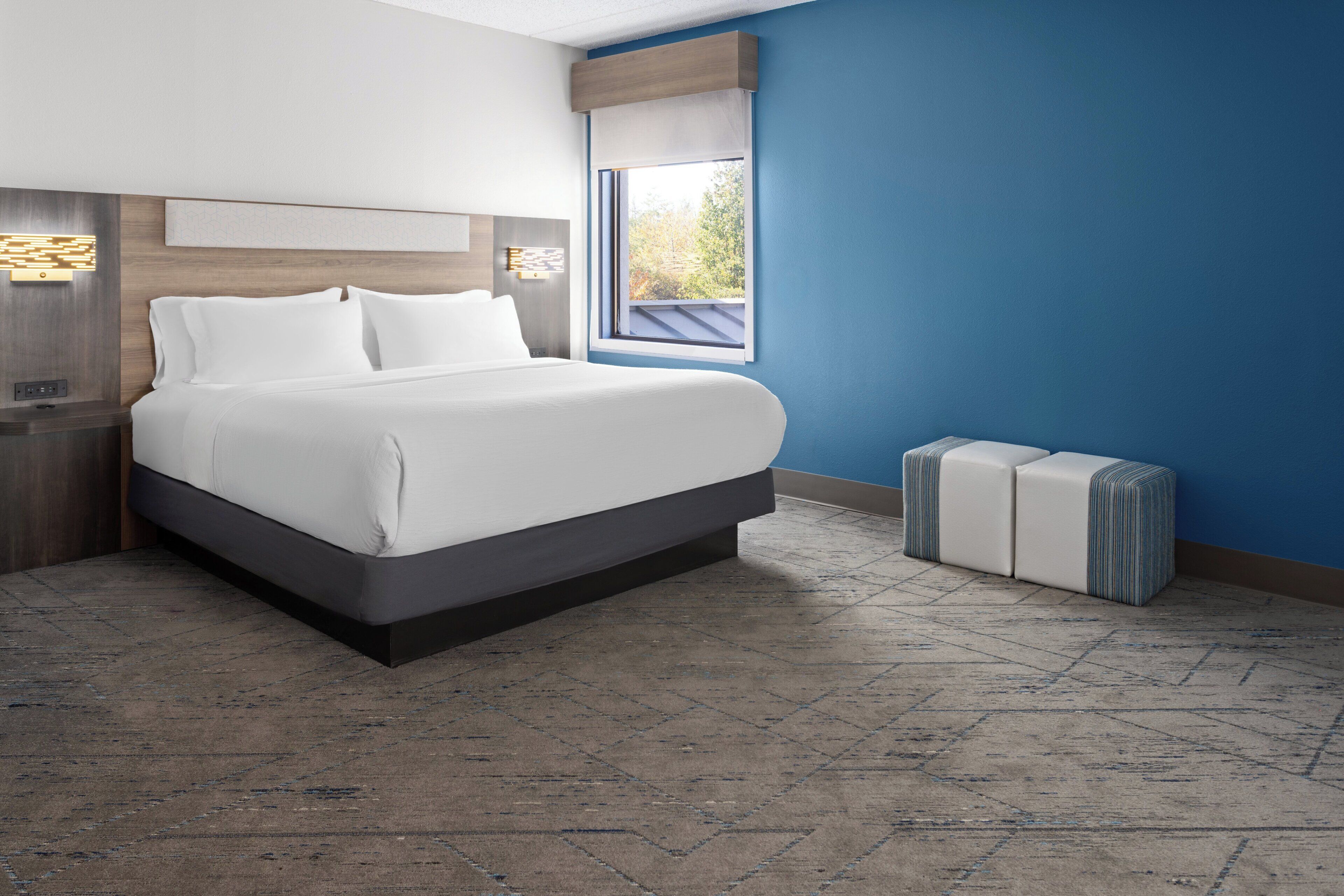 A modern hotel room with a large bed, a nightstand, a window with a view, and a built-in bench.