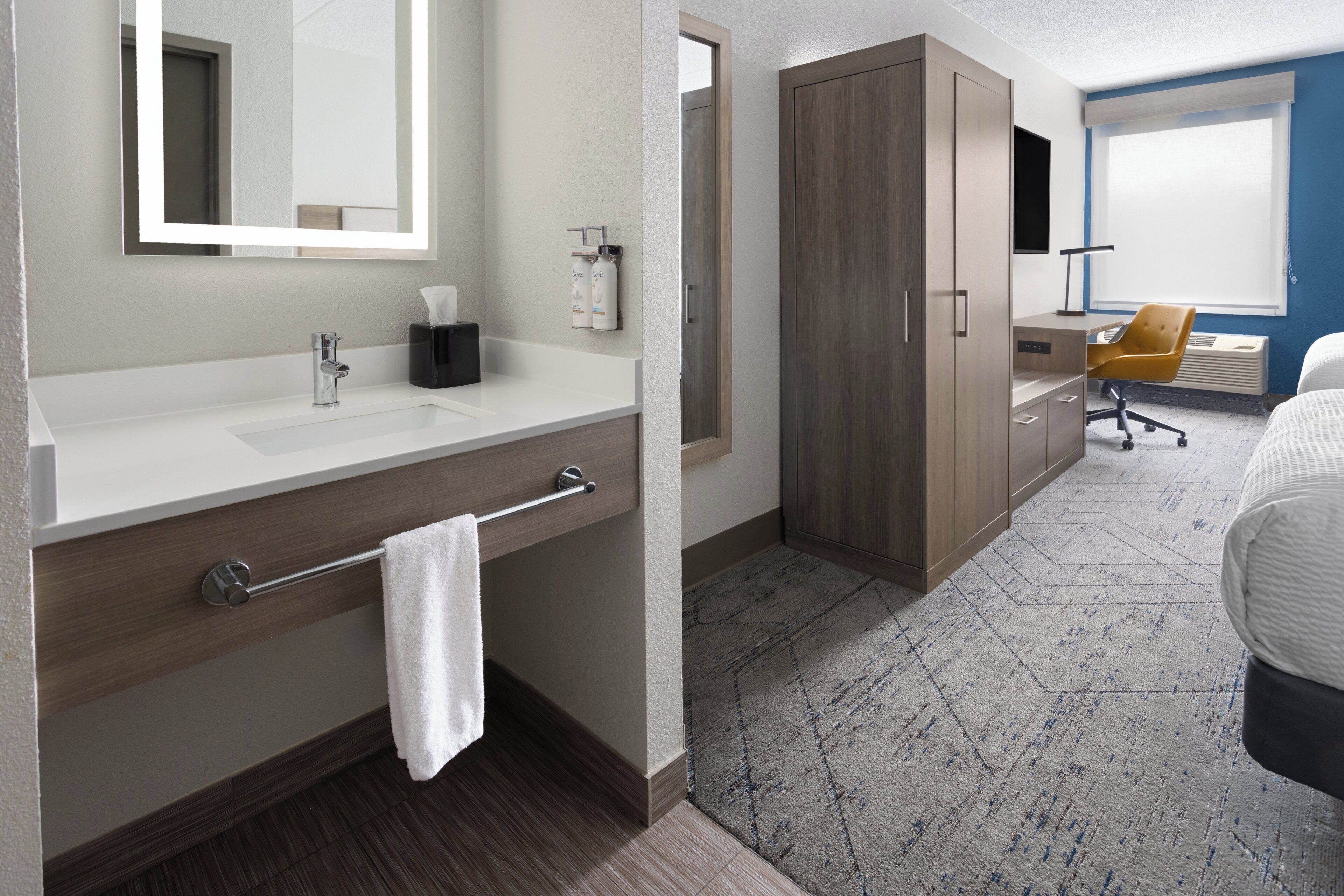 A modern hotel room with a white vanity, a large mirror, a wooden wardrobe, a desk with a chair, and a large bed.