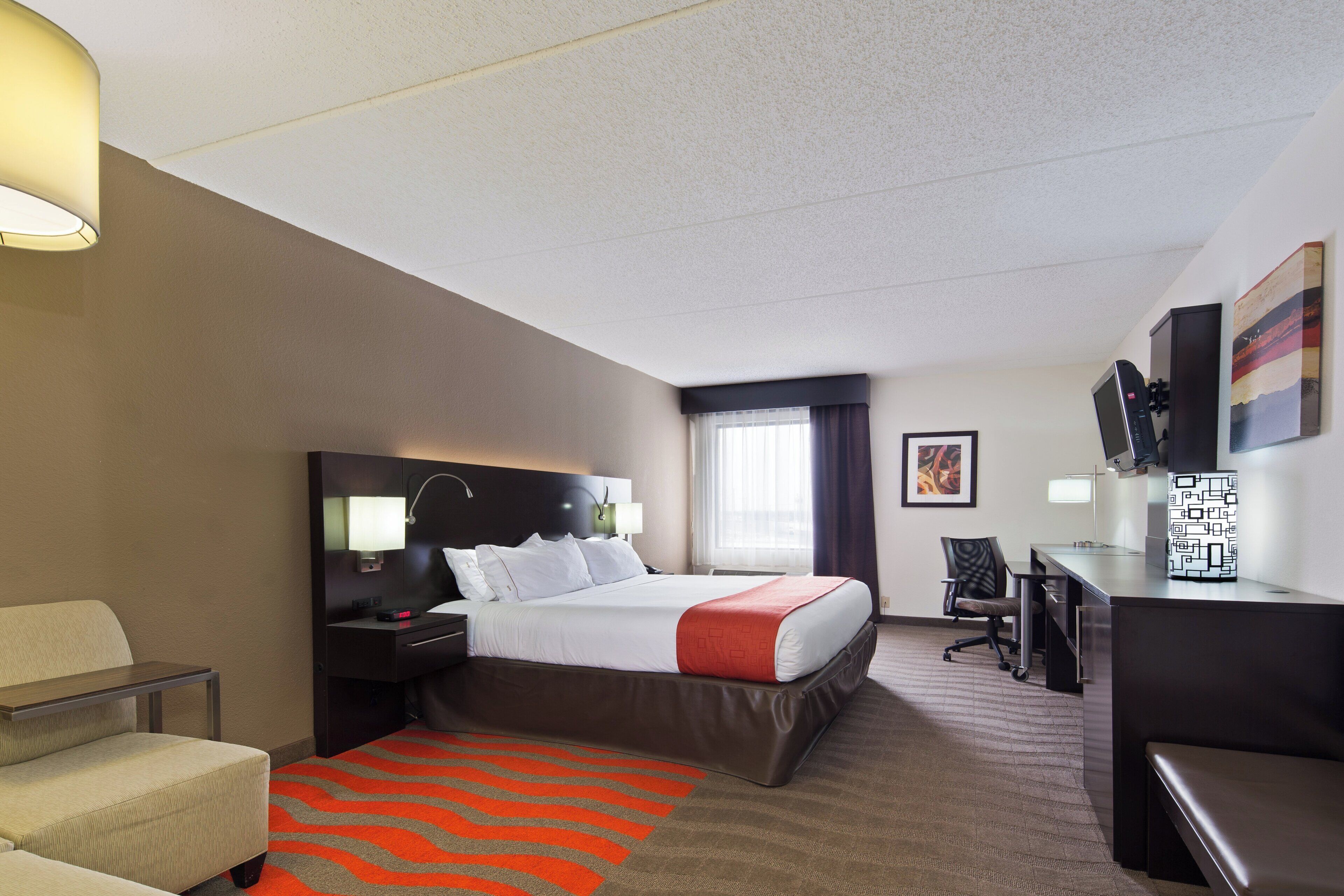 A hotel room with a bed, bedside table, lamp, desk, chair, and a view of the outdoors.
