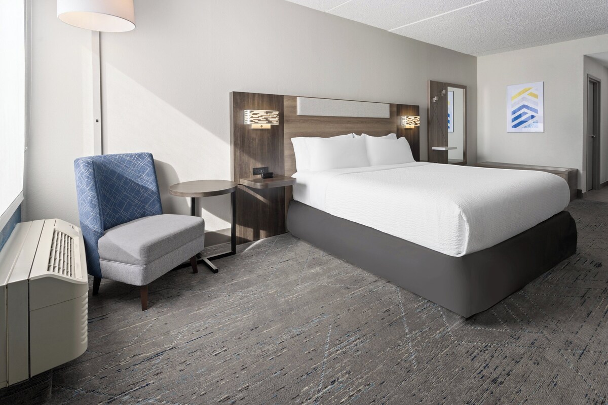 A hotel room with a large bed, a chair, a small table, and a lamp.