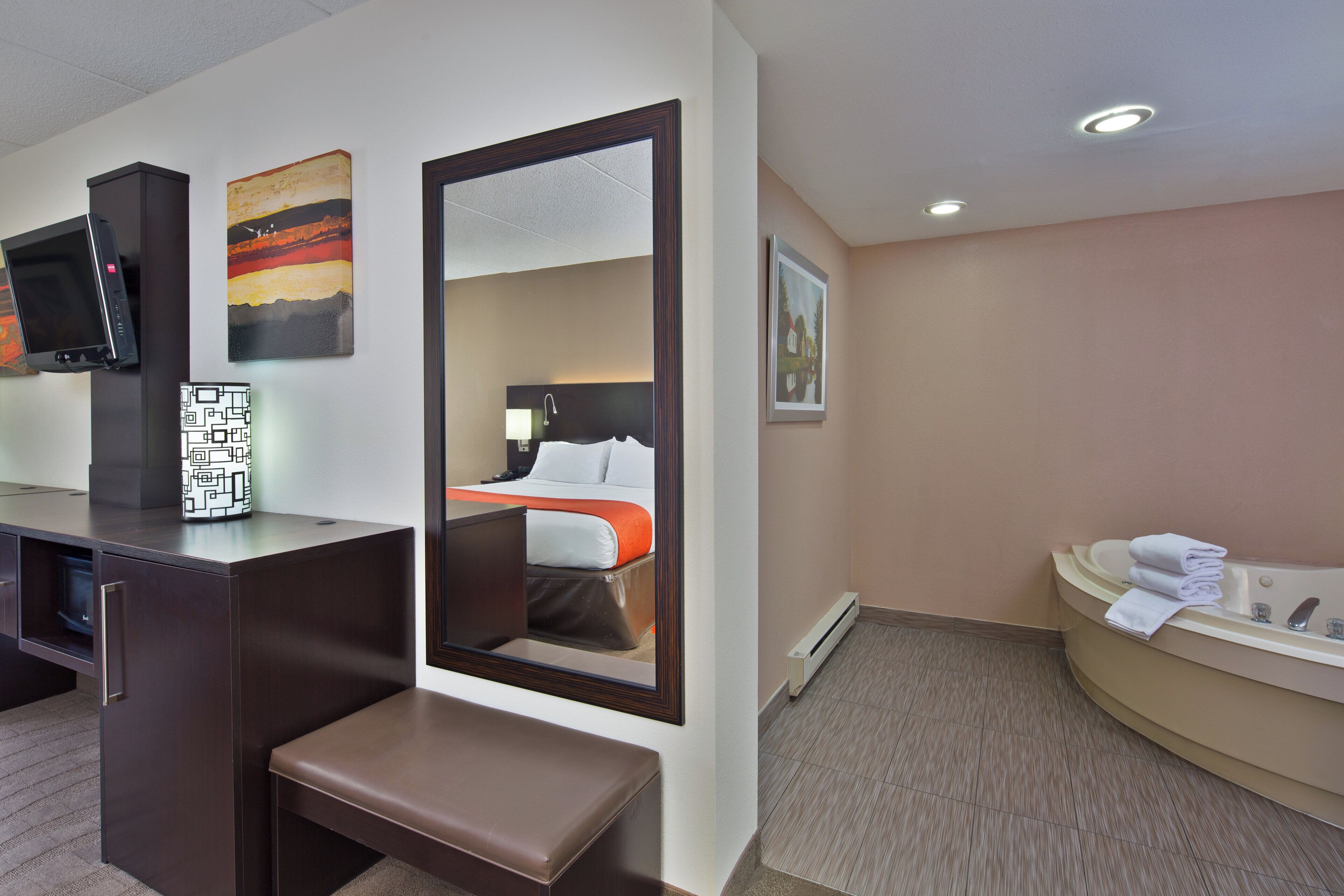 A hotel room with a bed, a desk, a bench, a TV, and a bathroom with a bathtub and towels.