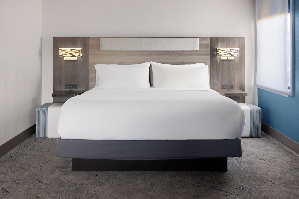 A modern hotel room with a large bed, two bedside tables with lamps, a headboard with a geometric light panel, and a view of the outdoors through a window.