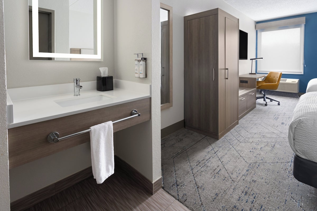 A modern hotel room with a white vanity, a large mirror, a wooden wardrobe, a desk with a chair, and a large bed.