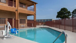 A swimming pool with a lifeguard stand and a blue chair, surrounded by a fenced area and a multi-story building with balconies.