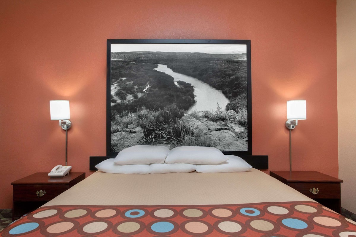 A hotel room with a bed, two bedside tables, a nightstand, a telephone, a pillow, a blanket, and a large framed picture of a river landscape on the wall.