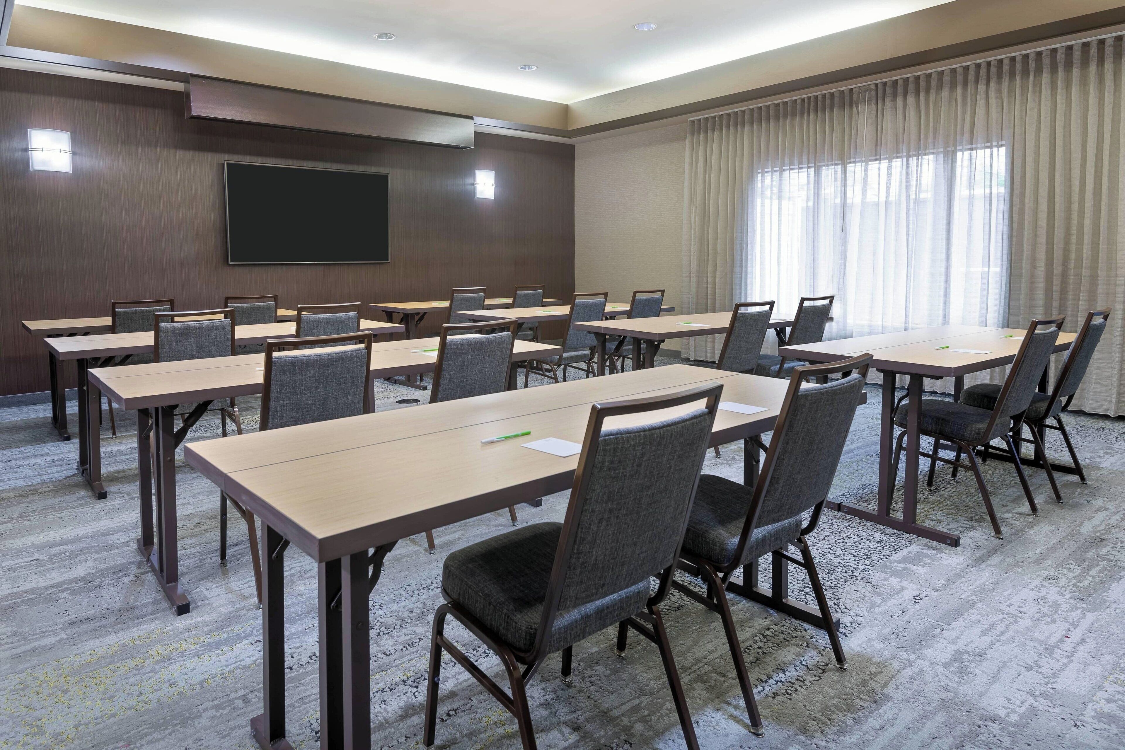 A conference room with multiple tables and chairs, a large screen, and a window with curtains.