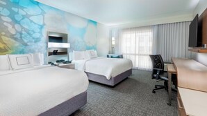 A hotel room with two beds, a desk, a chair, a TV, and a bathroom with a mirror and sink.