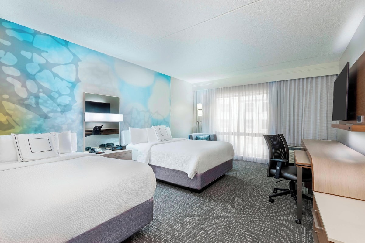 Une chambre d’hôtel avec deux lits, un bureau, une chaise, une télévision et une salle de bain équipée d’un miroir et d’un lavabo.
