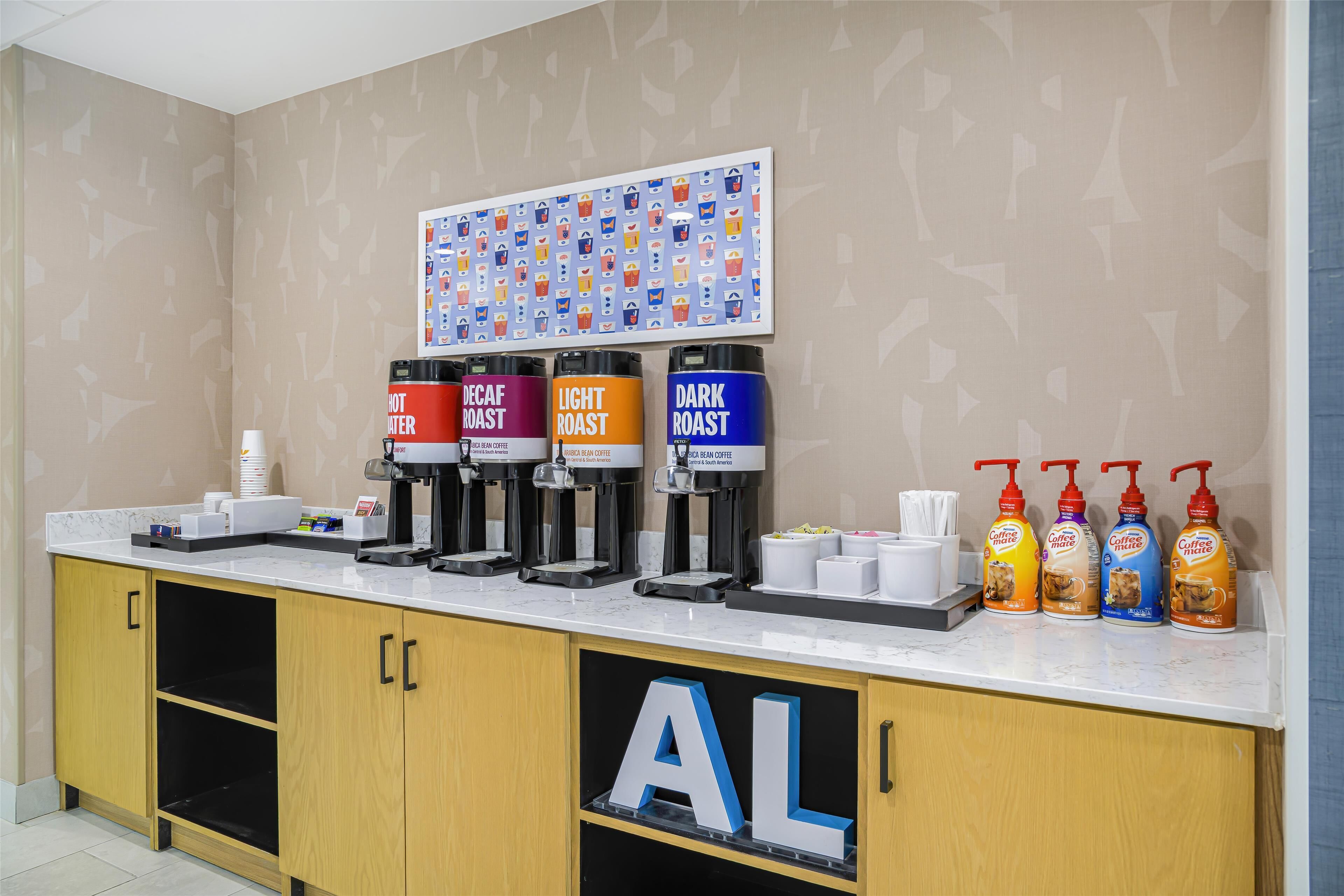 Estação de café com quatro dosadores identificados como 'Quente', 'Suave', 'Descafeinado' e 'Torra Escura'. Também há quatro garrafas de leite para café.