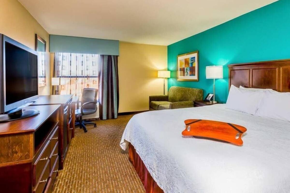 A hotel room with a bed, a TV, a desk, a chair, a lamp, and a painting on the wall.