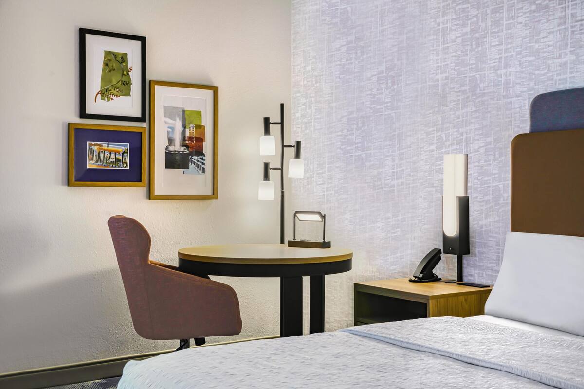 A hotel room with a bed, a desk, a chair, a small table, and framed pictures on the wall.
