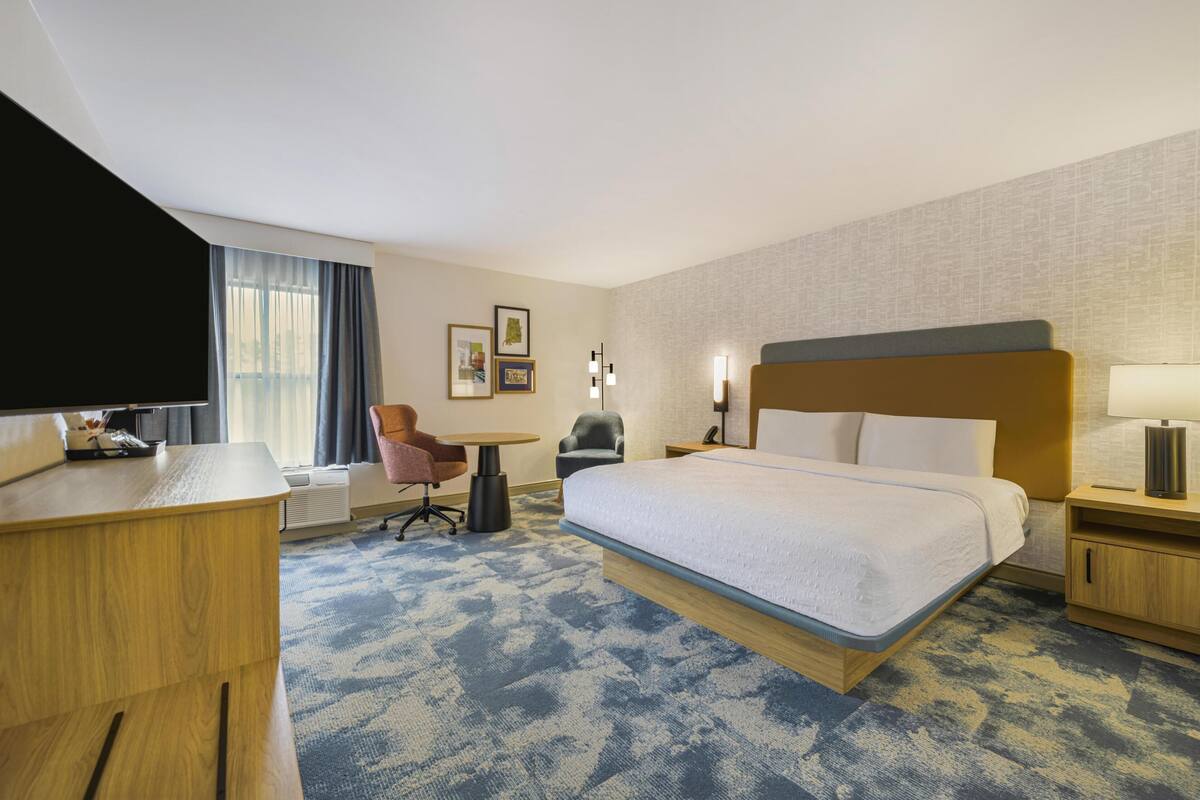 A hotel room with a large bed, a desk, a chair, a TV, and a window with curtains.