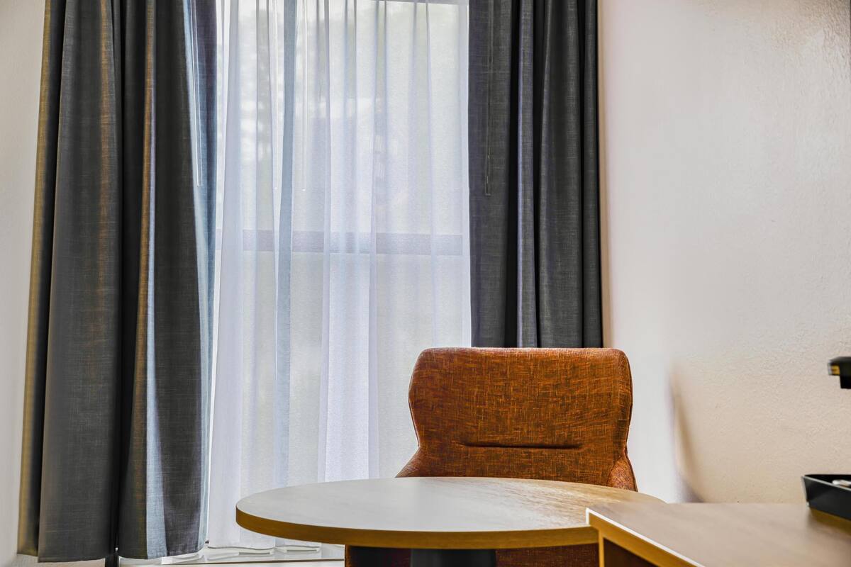 A room with a round table, a single orange chair, and a window with sheer curtains.