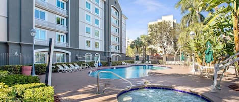 Zona de piscina de hotel con hidromasaje, sillas de descanso y un edificio con balcones.