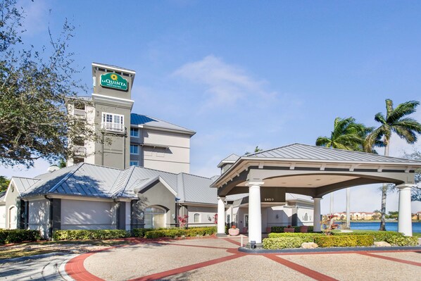 Entrada de hotel con zona de espera cubierta, palmeras y cielo azul.