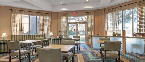 A hotel dining area with multiple tables and chairs, a bar, and large windows with curtains.