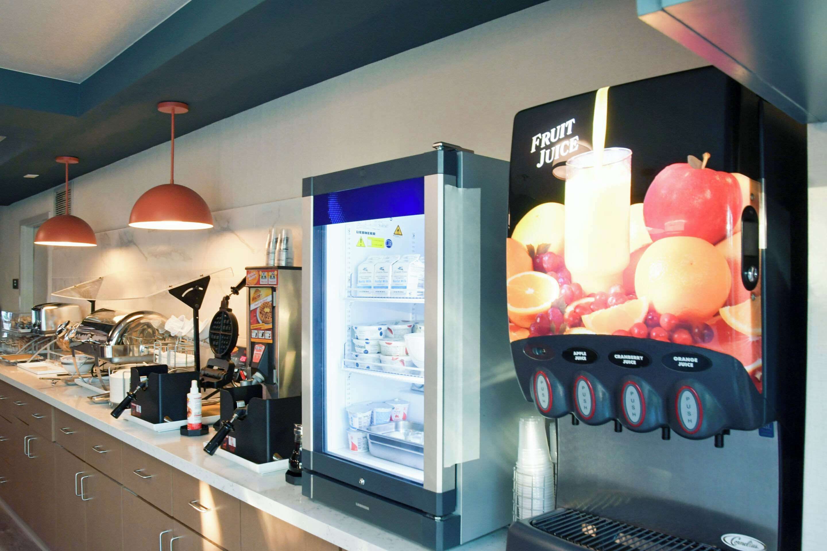 Una estación de bebidas autoservicio con múltiples dispensadores, un refrigerador y una zona de preparación de alimentos con una máquina para gofres.