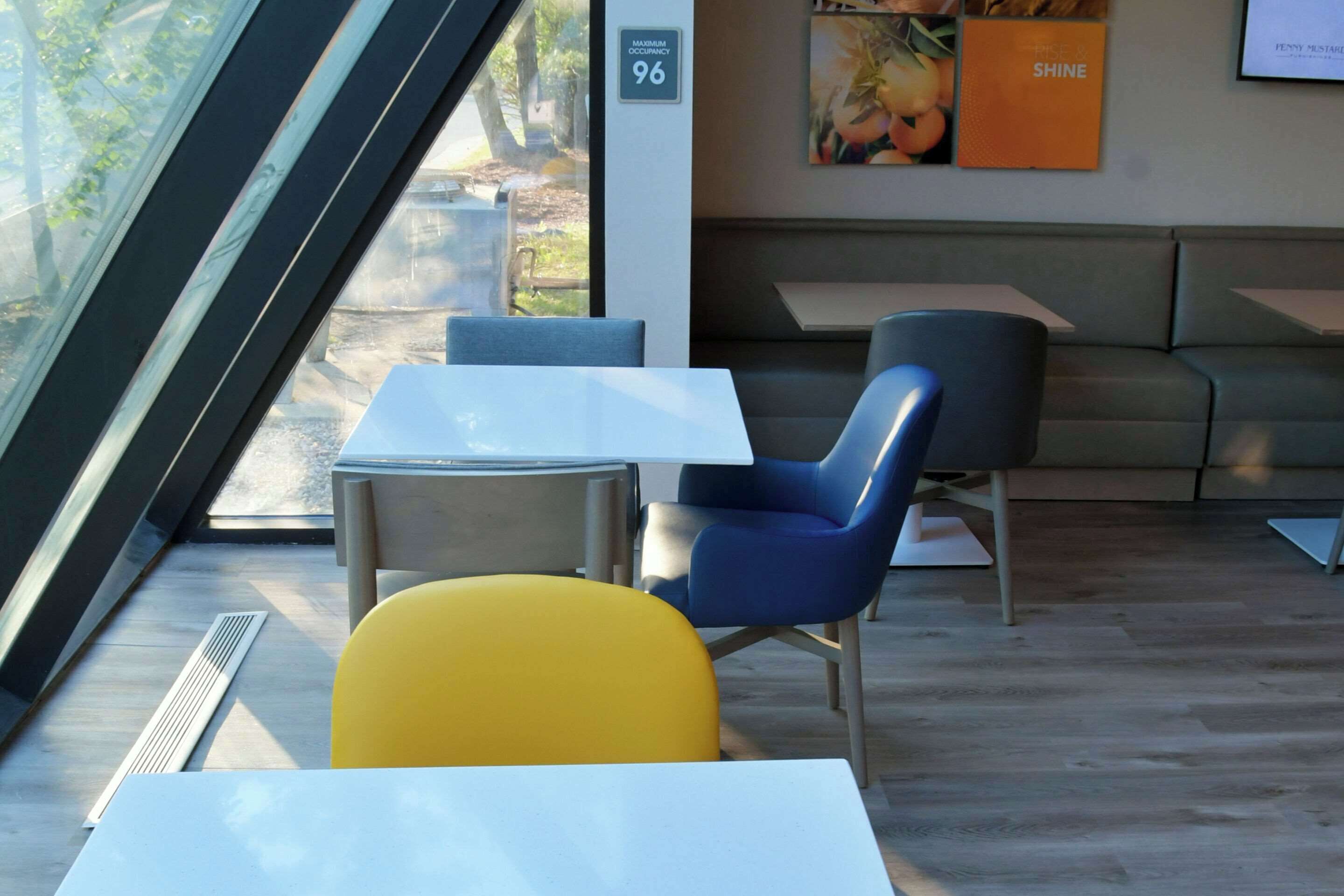 Un interior moderno con una zona de estar que incluye una silla amarilla, una silla azul y un sofá gris. Hay una mesa blanca y una rejilla de ventilación en el suelo. Una señal indica el número de habitación 96.