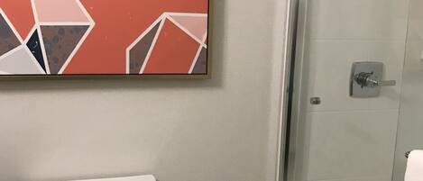 A bathroom with a toilet, a glass shower enclosure, and a geometric-patterned artwork on the wall.