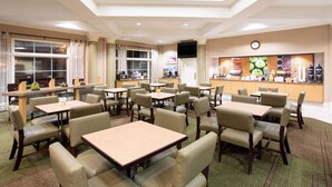 Zona de dinar d'hotel amb diverses taules i cadires, una barra amb aliments i begudes, i un televisor a la paret.