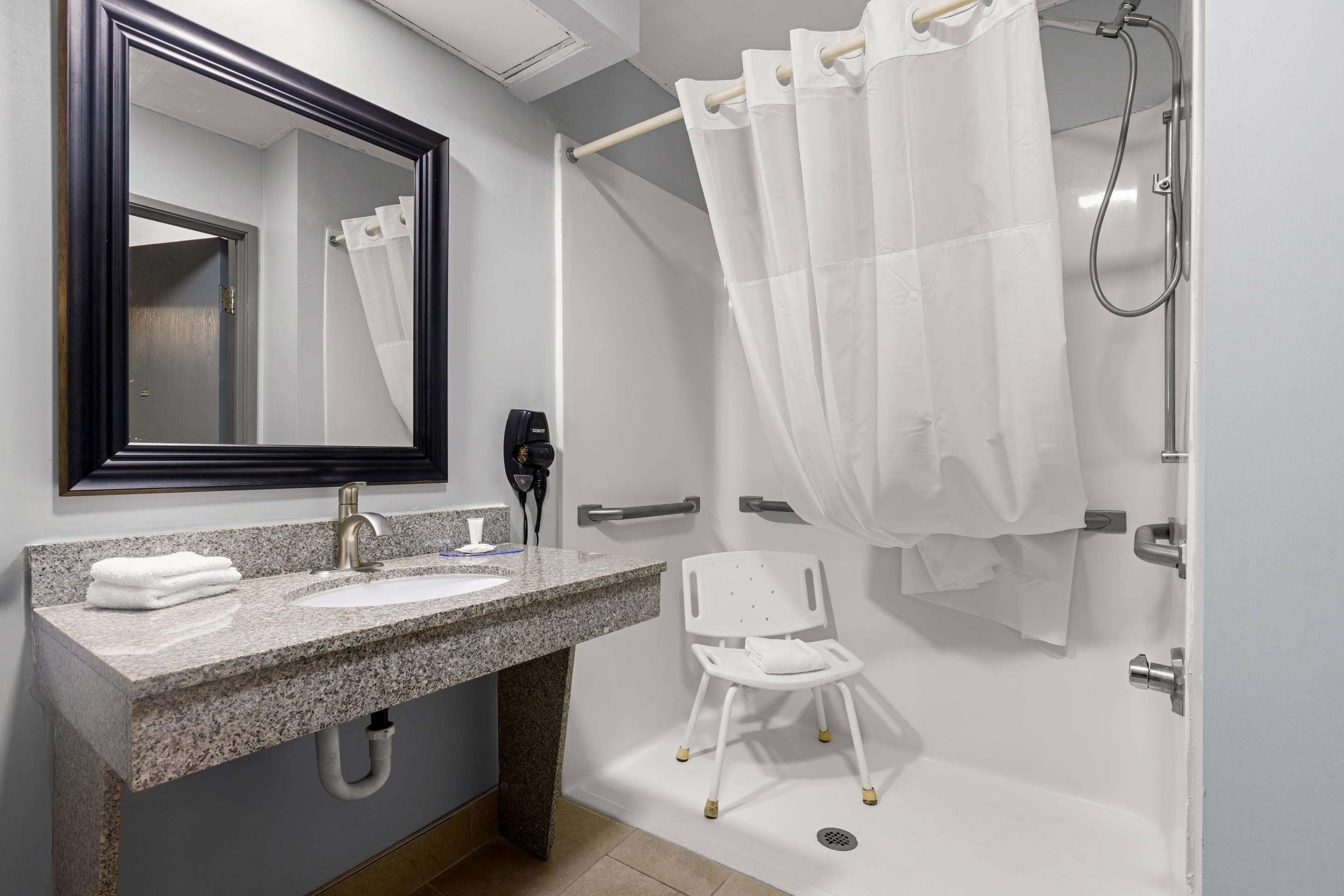 A modern bathroom with a granite countertop, a sink, a mirror, a shower with a white curtain, and a white chair.