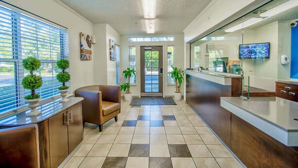 A reception area with a check-in counter, a seating area with a brown sofa, potted plants, and a tiled floor. -  Studio 6 Deer Valley (Phoenix)