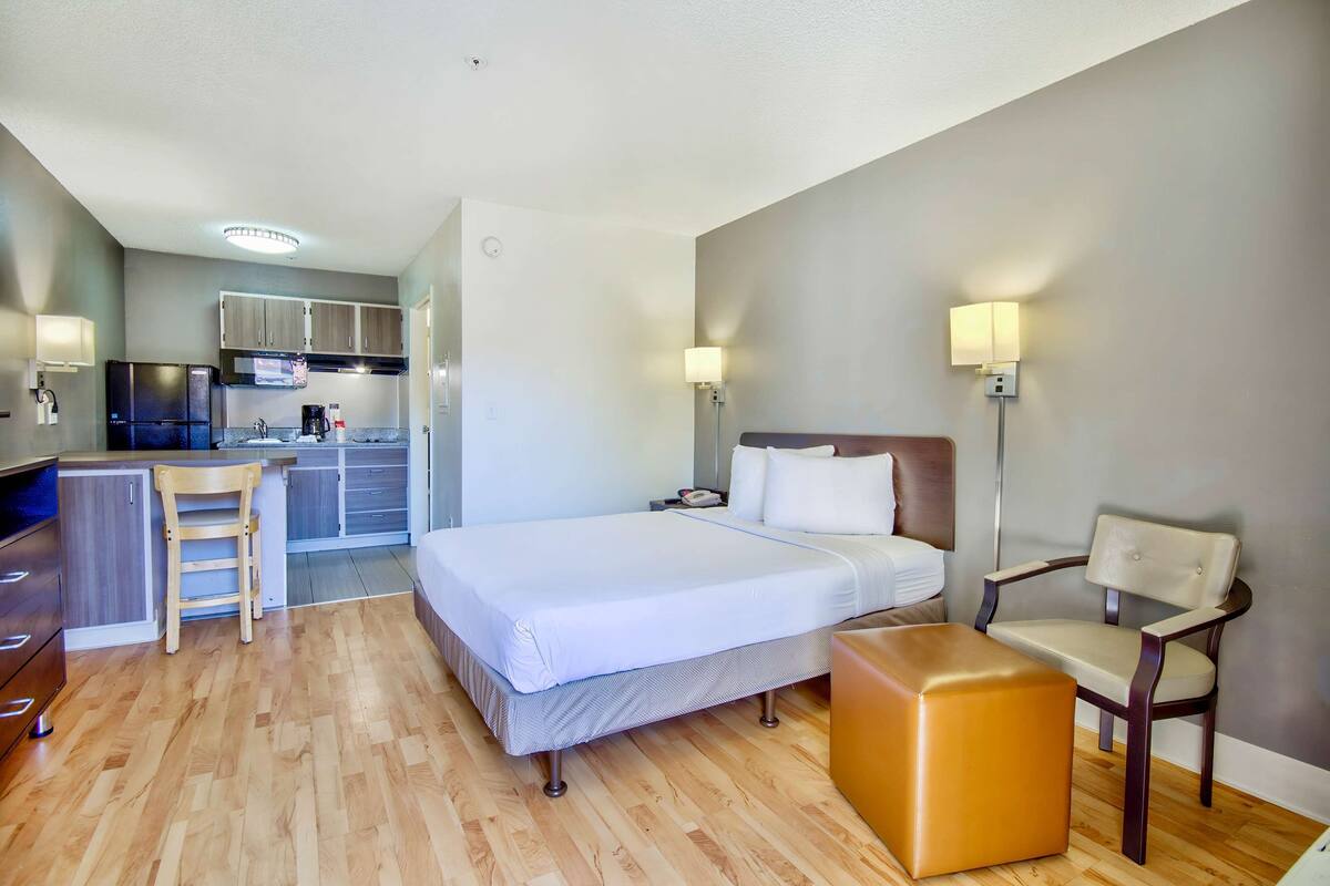 A hotel room with a bed, a desk, a chair, a small table, and a kitchenette in the background.
