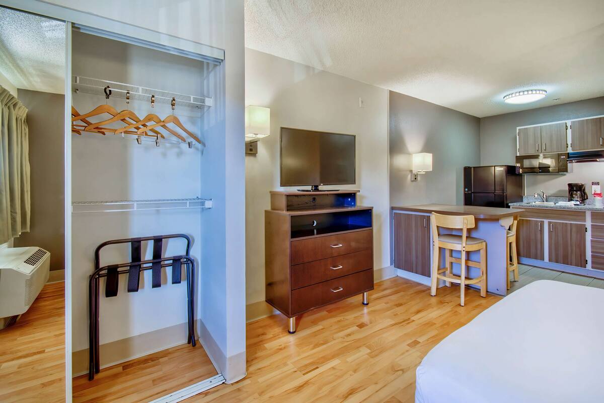 A hotel room with a wooden floor, a bed, a desk with a chair, a kitchenette, a TV, and a wardrobe with hangers.