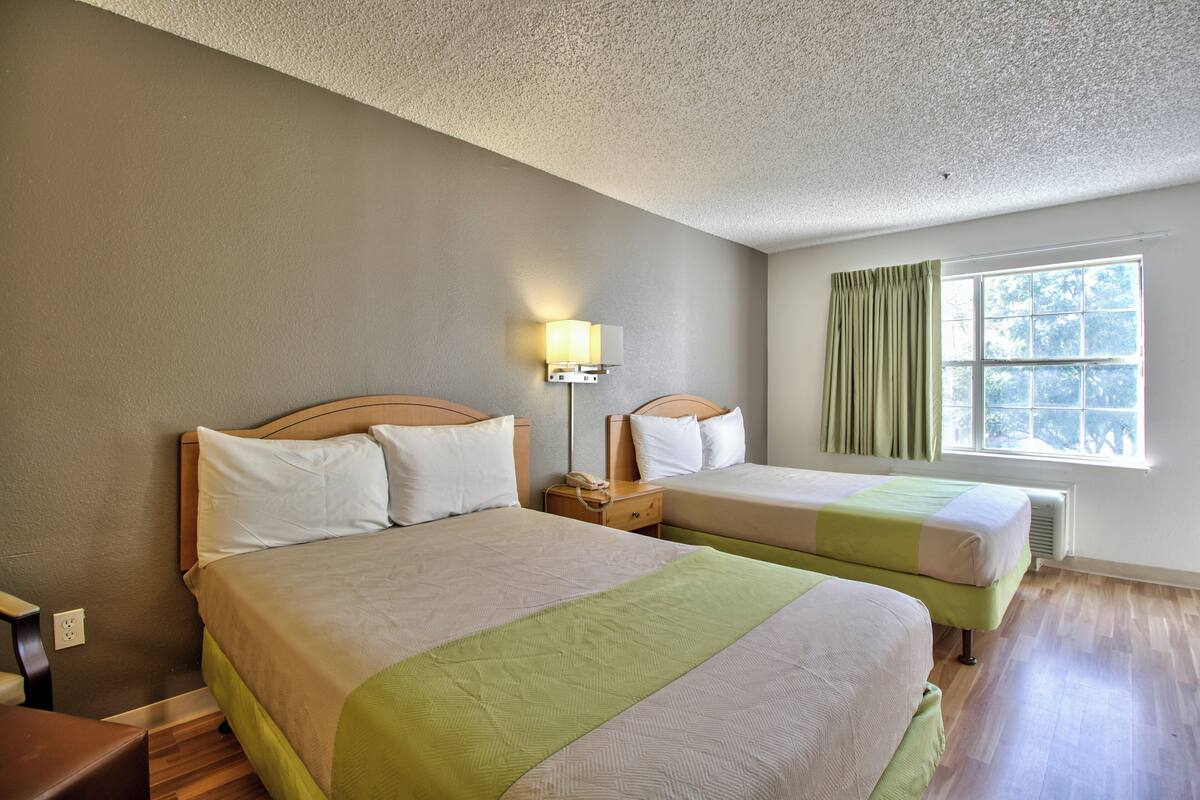 A hotel room with two beds, a nightstand with a lamp, a window with curtains, and a wooden floor.