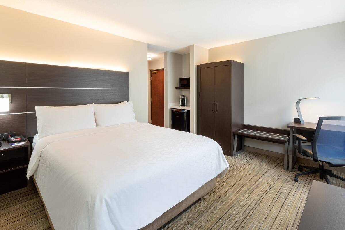 Une chambre d’hôtel avec un grand lit, un bureau avec une chaise, une armoire et une table de chevet avec une lampe.