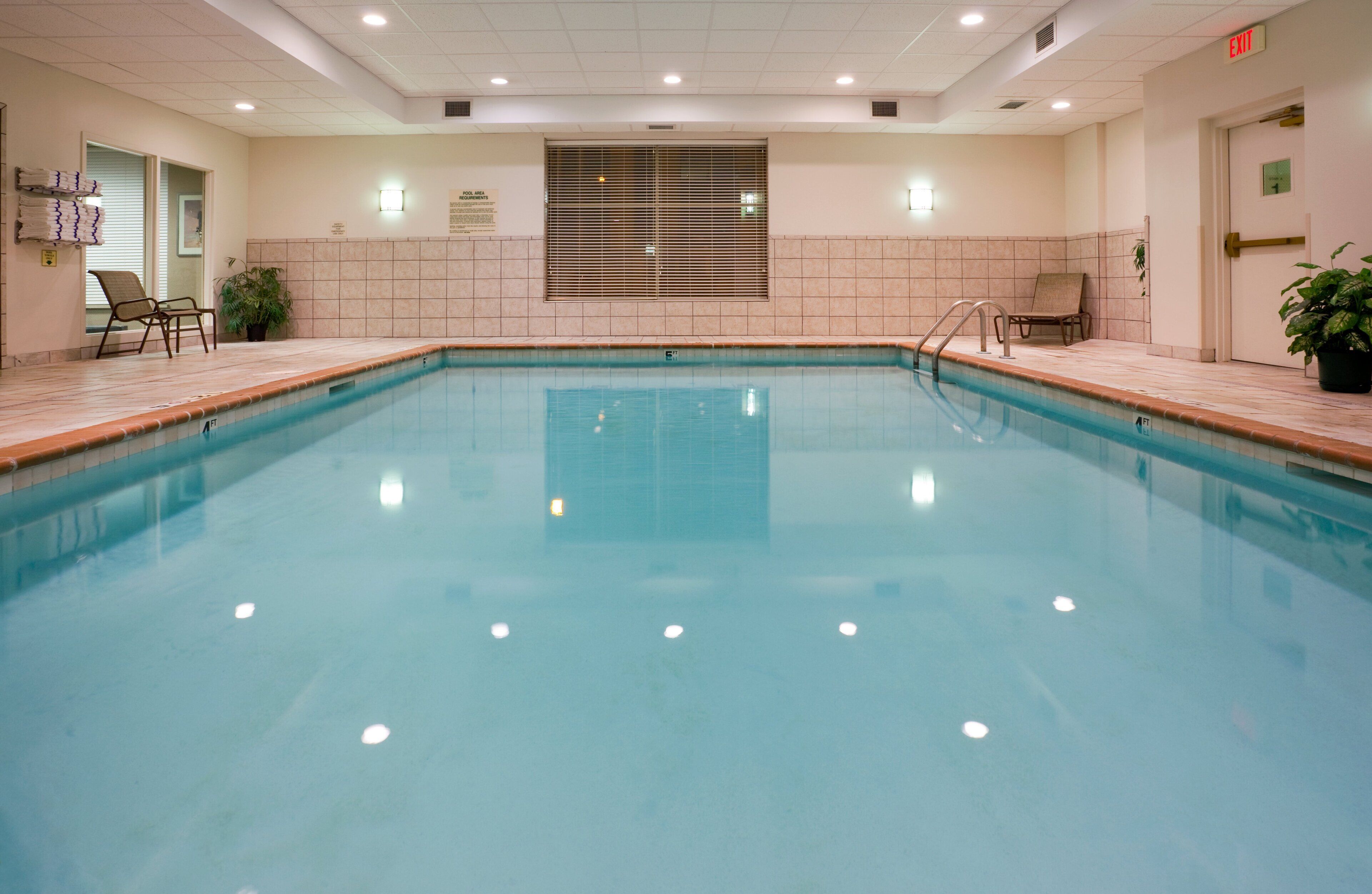 A clear indoor swimming pool with tiled walls, a window with blinds, and a few lounge chairs.