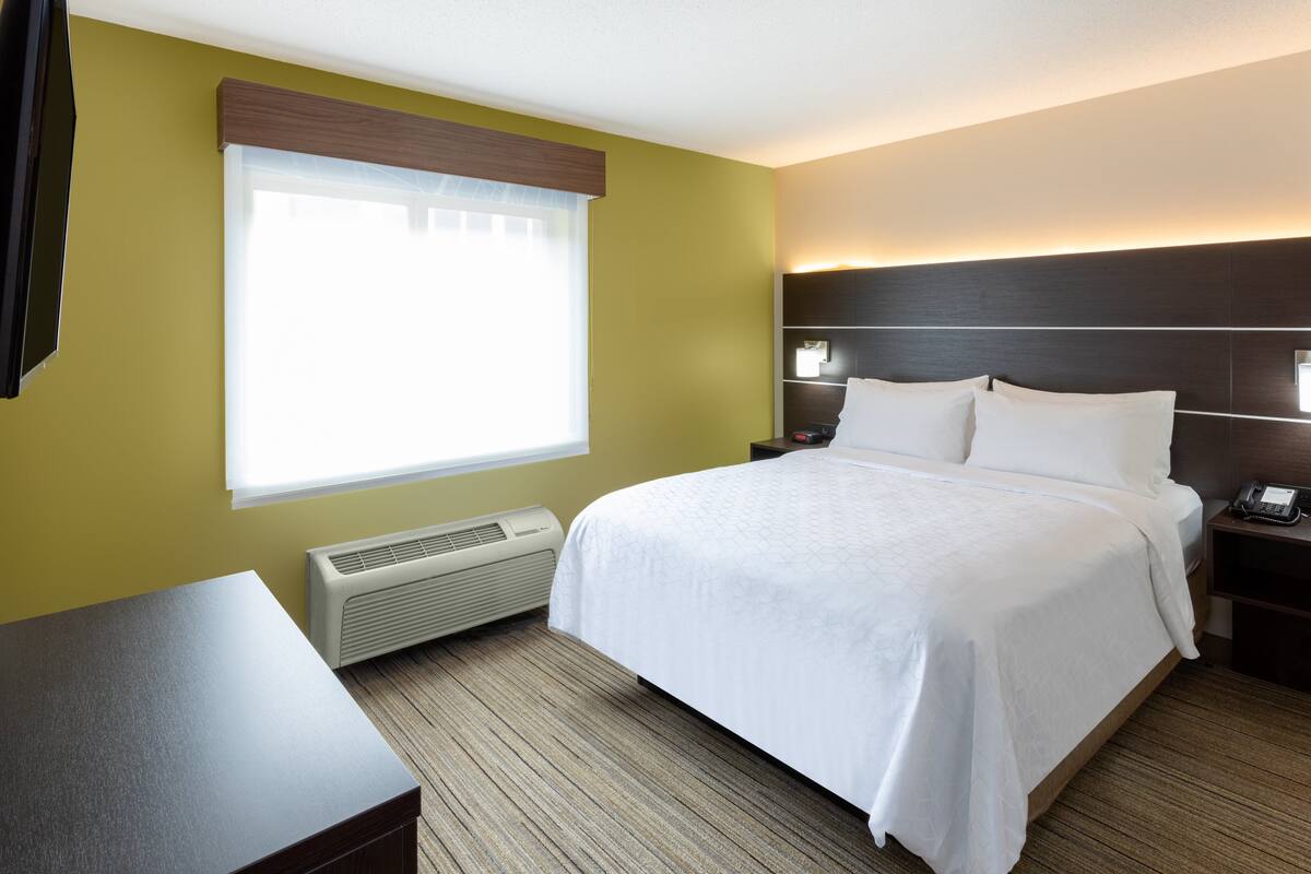 Une chambre d’hôtel comprenant un lit, des tables de chevet, un bureau, une fenêtre avec des stores et un radiateur fixé au mur.