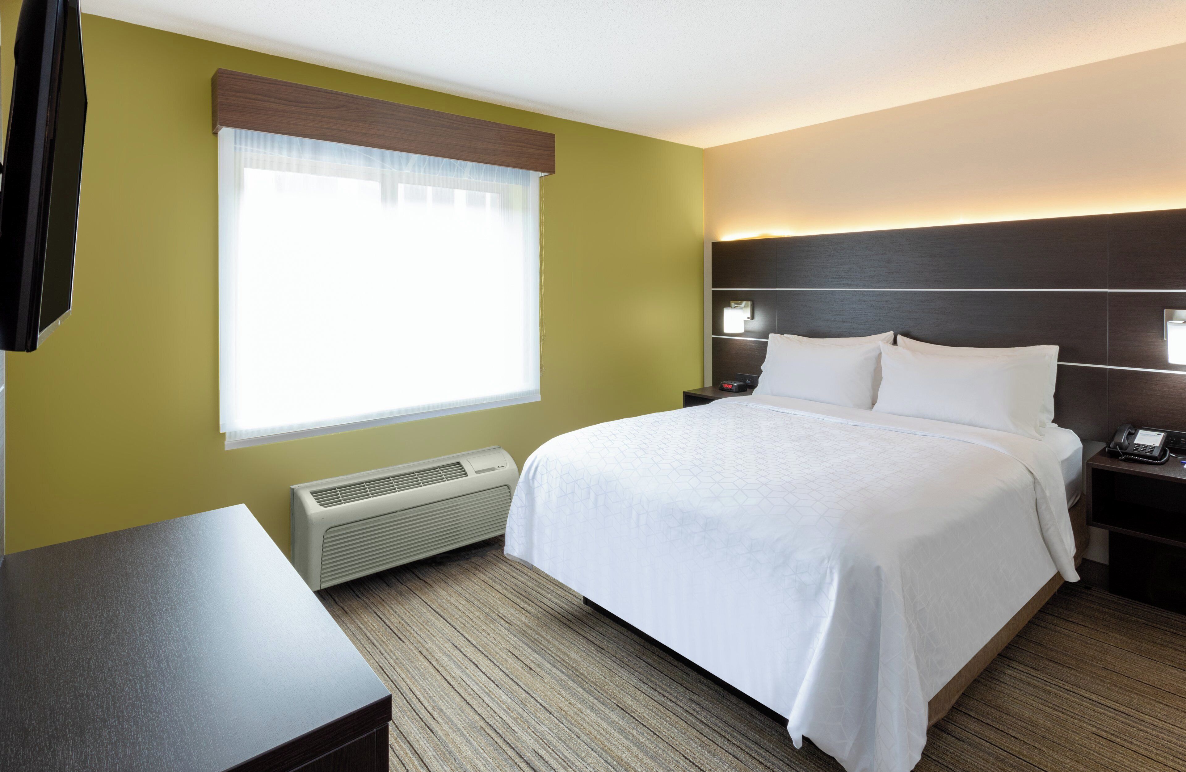 Une chambre d’hôtel comprenant un lit, des tables de chevet, un bureau, une fenêtre avec stores, un radiateur fixé au mur et une petite table de nuit avec un téléphone.
