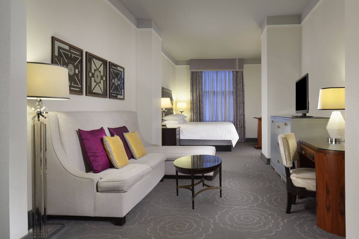 A hotel room with a sofa, a small table, a desk with a chair, a bed, a TV, and a window with curtains.